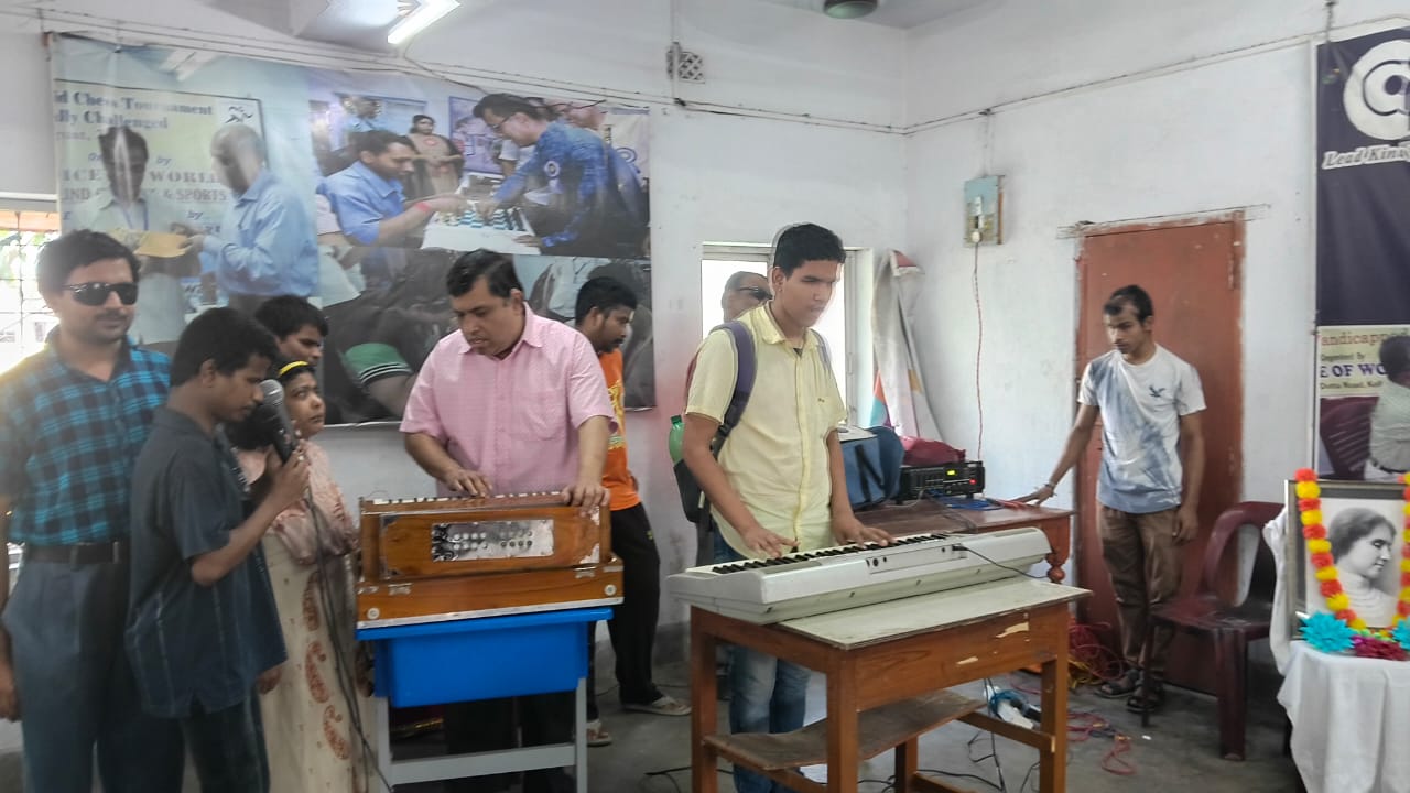 Students performing on harmonium and keyboard at the Helen Keller birthday celebration at Voice of World, with a garlanded portrait of Helen Keller in the background